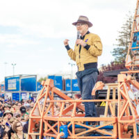 Adam Savage giving a speech while standing on the Electric Giraffe in front of a large crowd.