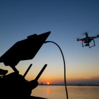 silhouette of airborne drone and pilot's controller against setting sun over ocean.