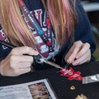 a young woman sitting at a table soldering the legs of an LED on the back of a small rocket shaped wearable pin